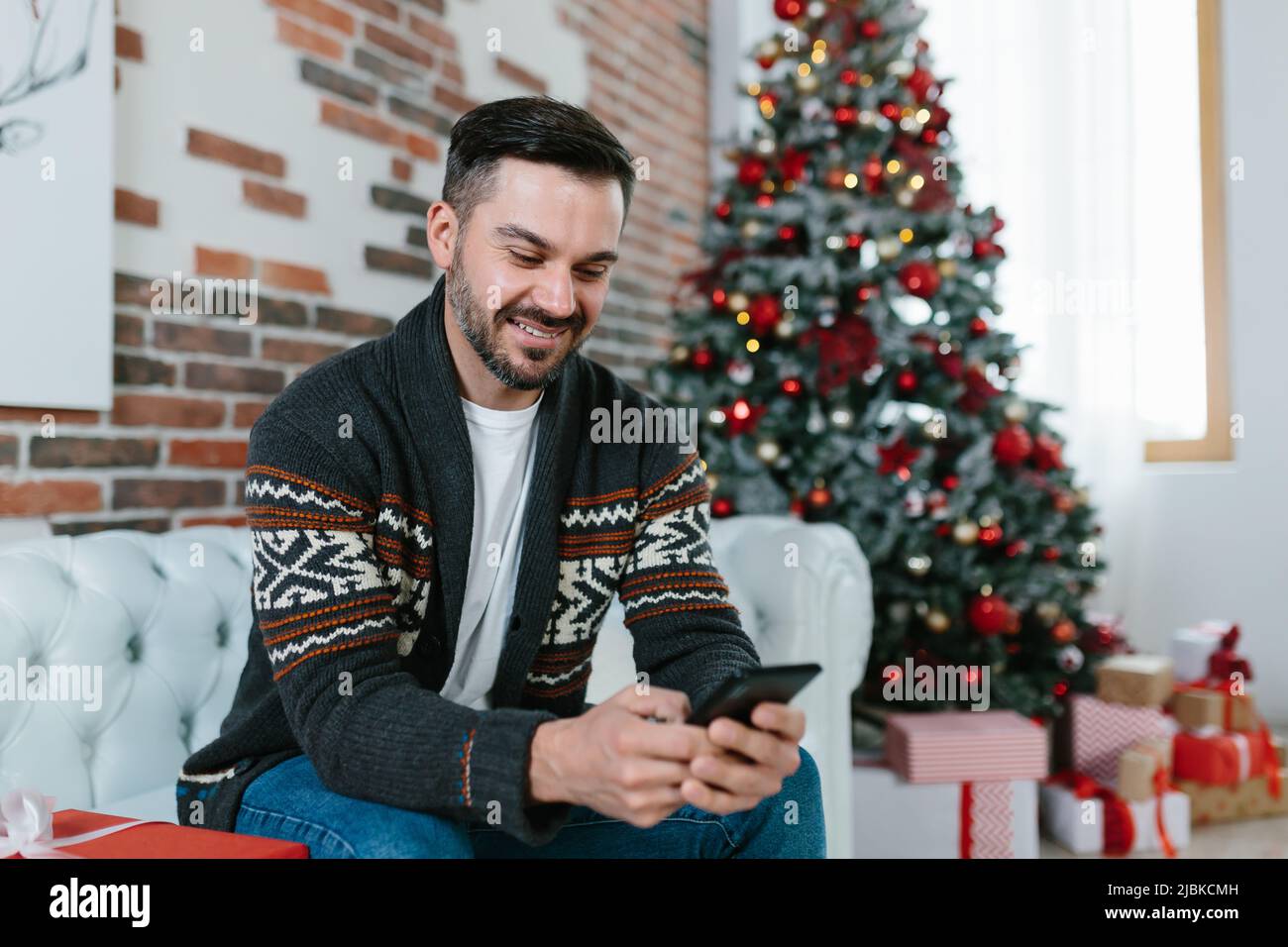 Glücklicher Mann, der zu Hause auf der Couch sitzt, korrespondiert auf einem Handy mit Freunden, schaut auf das Telefon in der Nähe des Weihnachtsbaums und wartet auf die Stockfoto