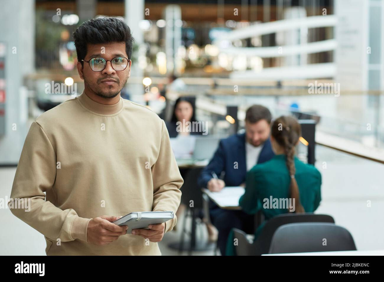 Waist-up-Porträt eines jungen Mannes indischer Herkunft, der einen Planer hält und die Kamera im Bürogebäude anschaut, Raum kopieren Stockfoto