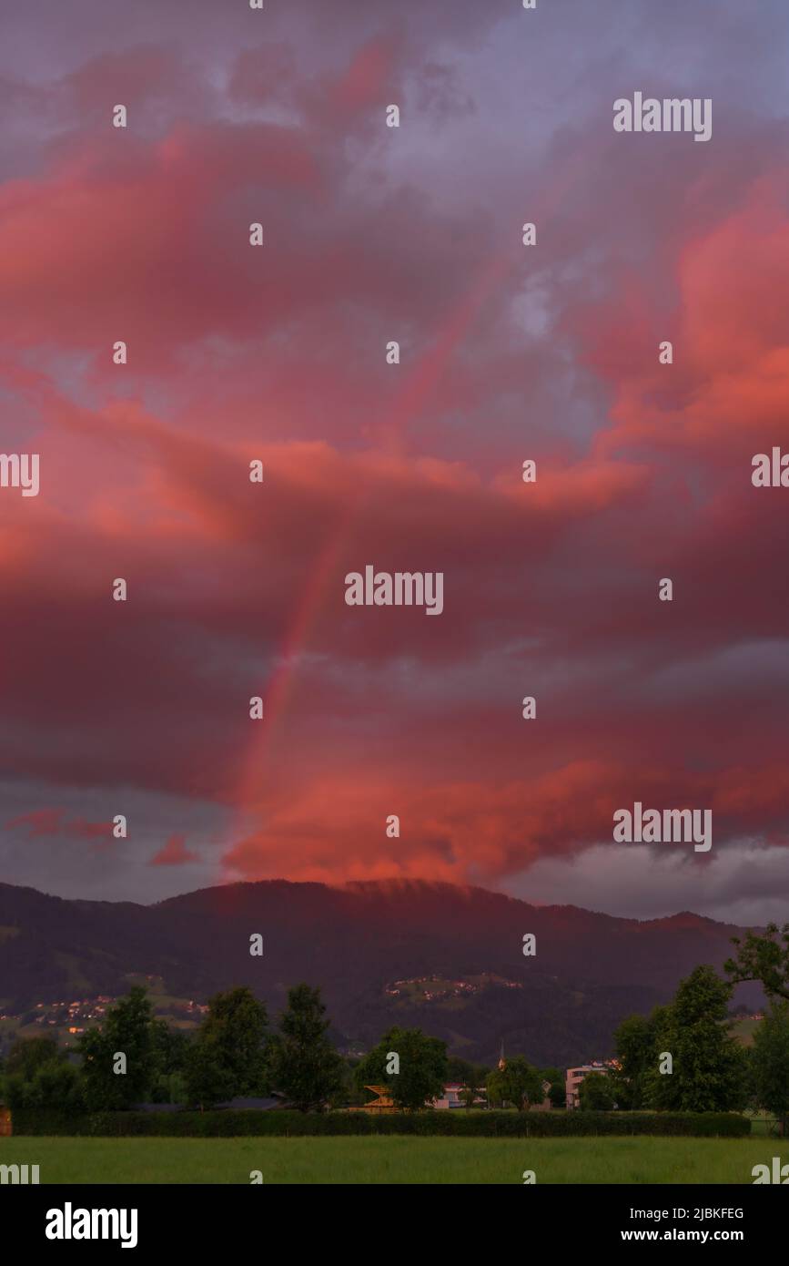 leuchtendes Abendrot mit Regenboden nach dem Sonnenuntergang in Dornbirn, Vorarlberg, Österreich. rote und graublaue Wolken über den grünen Wiesen Stockfoto