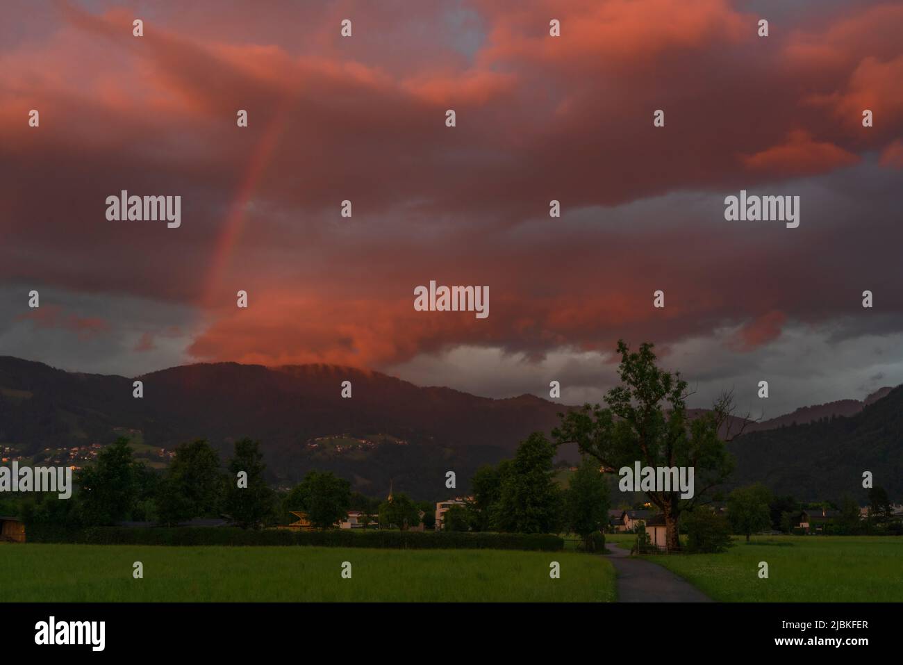 leuchtendes Abendrot mit Regenboden nach dem Sonnenuntergang in Dornbirn, Vorarlberg, Österreich. rote und graublaue Wolken über den grünen Wiesen Stockfoto