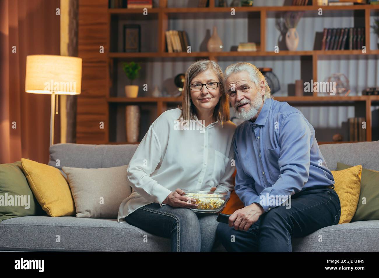 Seniorehepaar, grauhaariger Mann und Frau, sitzen zu Hause und sehen fern, essen abends Popcorn, schauen auf die Kamera und lächeln Stockfoto