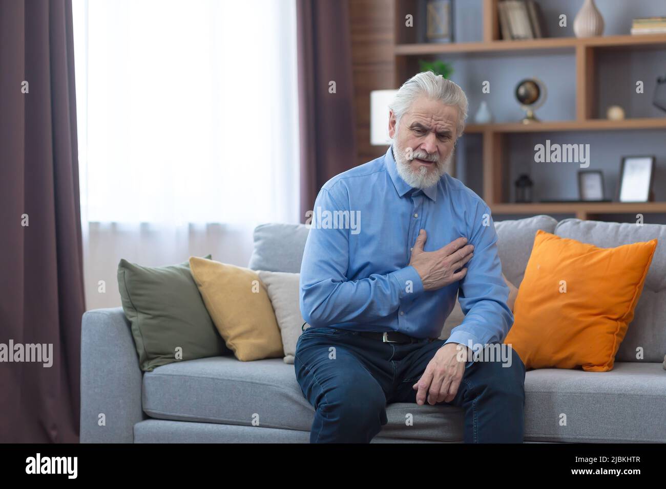 Allein ein älterer Mann zu Hause, der auf dem Sofa sitzt, die Hände auf der Brust hält, hat starke Brustschmerzen Stockfoto
