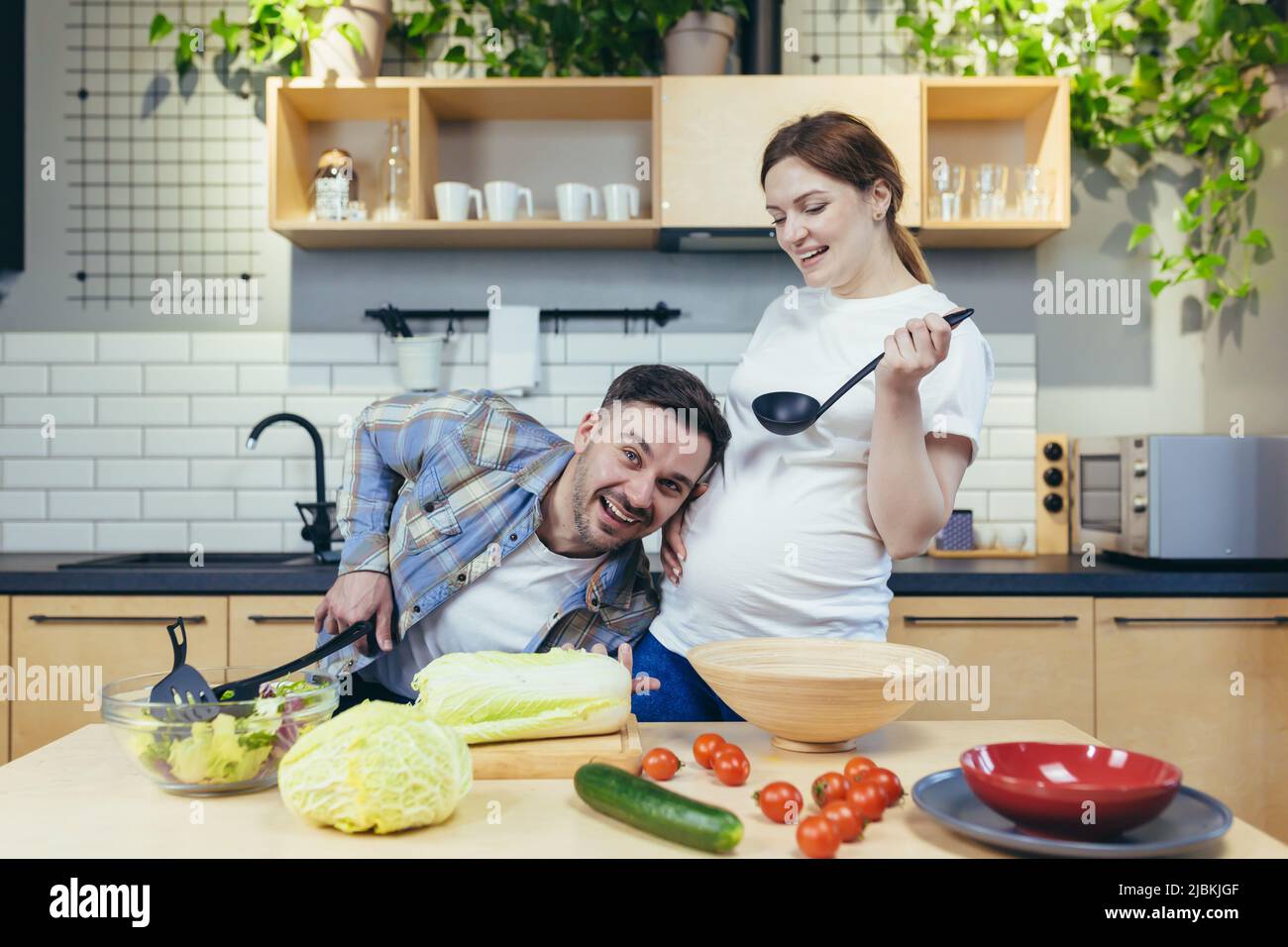 Eine junge Familie, eine Schwangere und ein Mann bereiten gemeinsam in der Küche gesundes und leckeres Essen zu. Viel Spaß zusammen. Der Mann wird seinem wi helfen Stockfoto