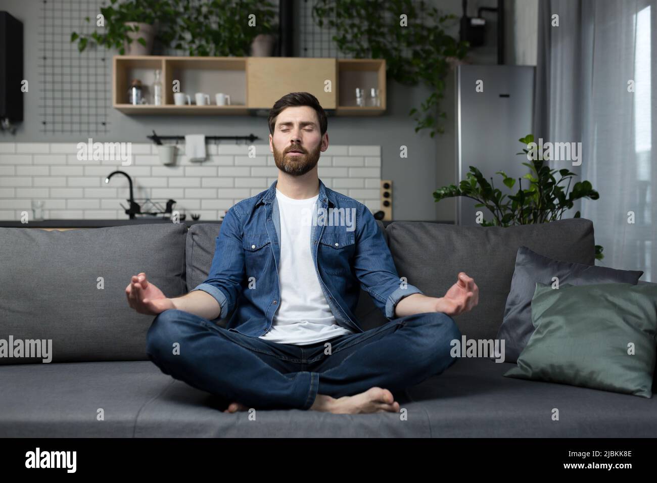 Junger Mann mit einem Bart allein zu Hause, sitzt auf der Couch und meditiert in der Lotusposition, in lässiger Kleidung Stockfoto