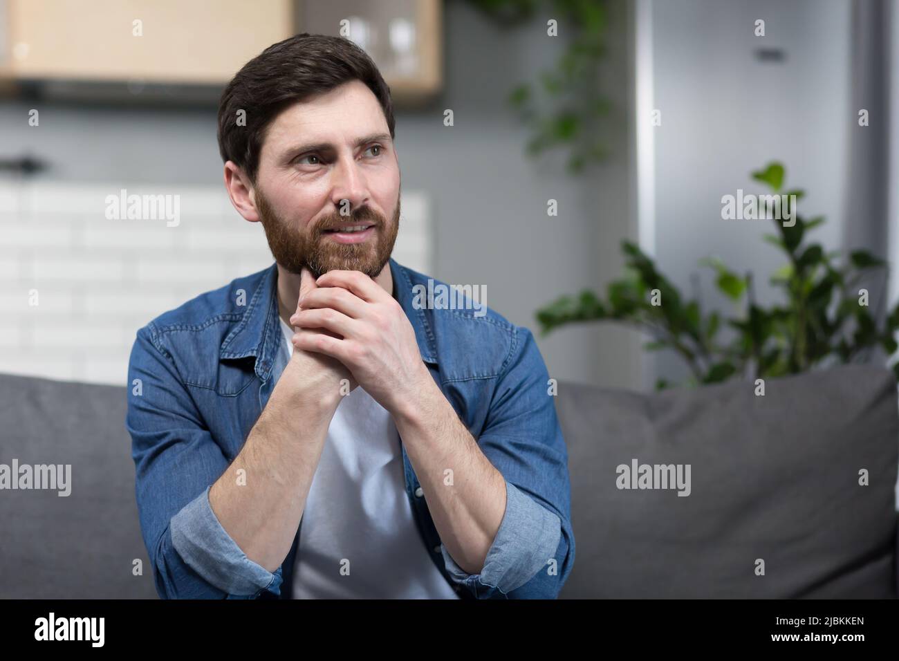 Der Mann zu Hause sitzt auf der Couch, ruht sich aus und schaut weg, lächelt verträumt nachdenklich Stockfoto
