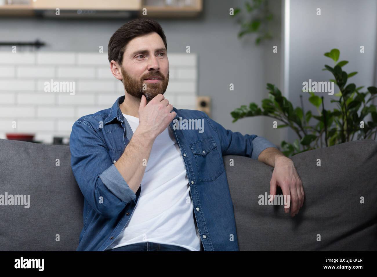 Der Mann zu Hause sitzt auf der Couch, ruht sich aus und schaut weg, lächelt verträumt nachdenklich Stockfoto