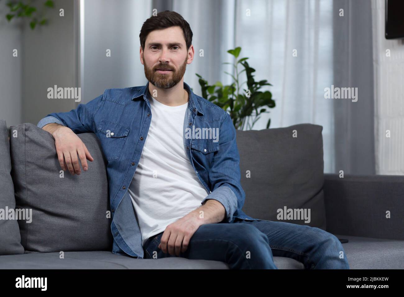 Porträt eines Mannes mit Bart zu Hause, der auf der Couch sitzt und die Kamera anschaut Stockfoto