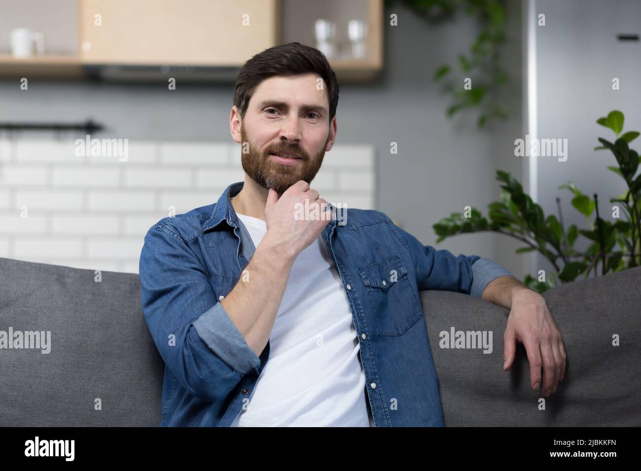 Porträt eines Mannes mit Bart zu Hause, der auf der Couch sitzt und die Kamera anschaut Stockfoto