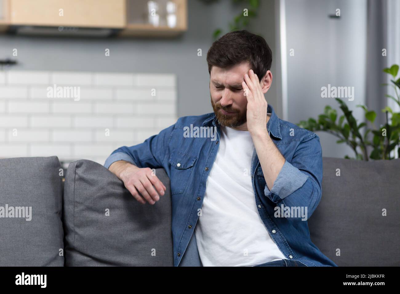 Der Mann ist zu Hause, der Patient sitzt auf der Couch, hat starke Kopfschmerzen Stockfoto