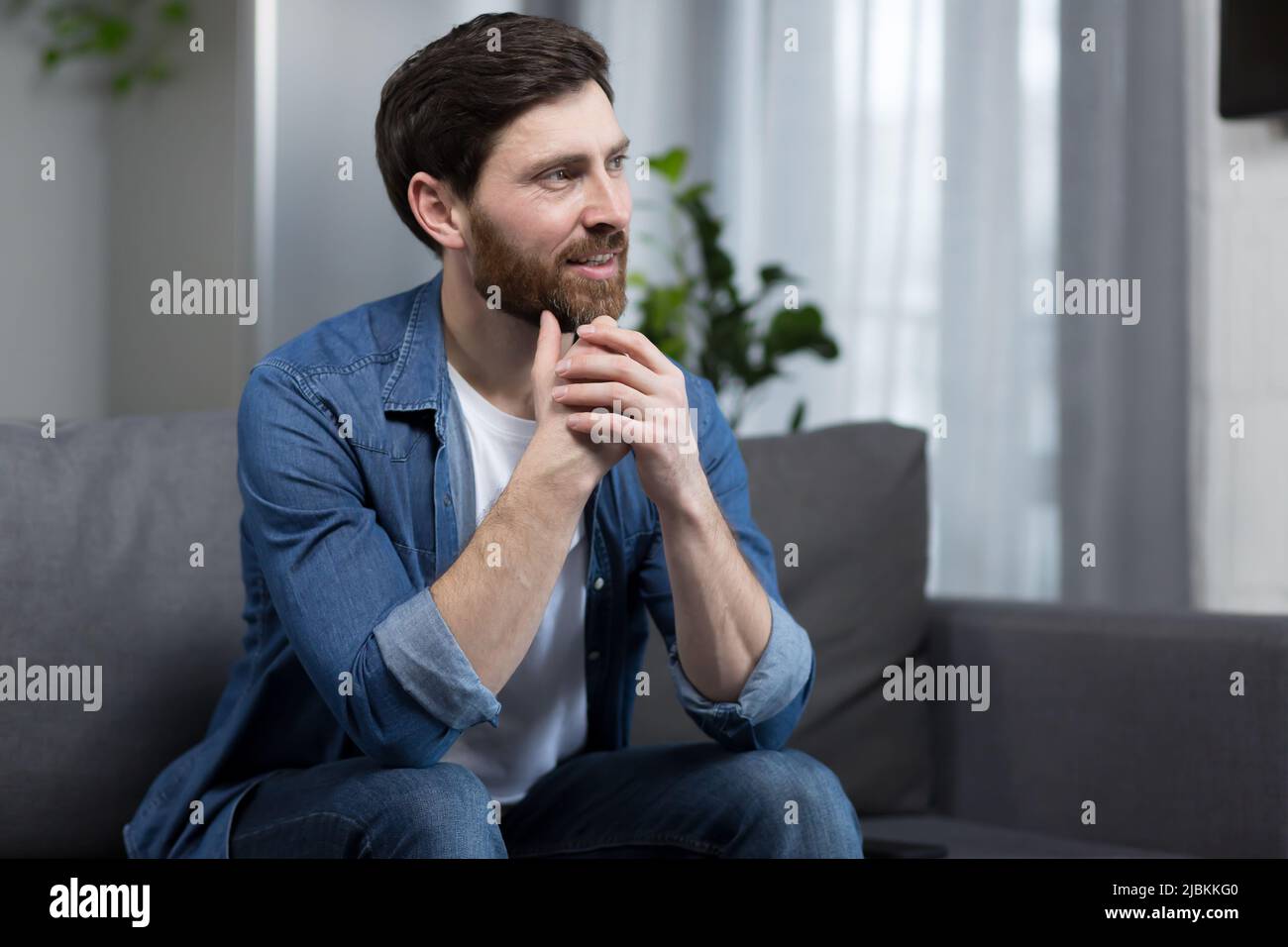 Der Mann zu Hause sitzt auf der Couch, ruht sich aus und schaut weg, lächelt verträumt nachdenklich Stockfoto