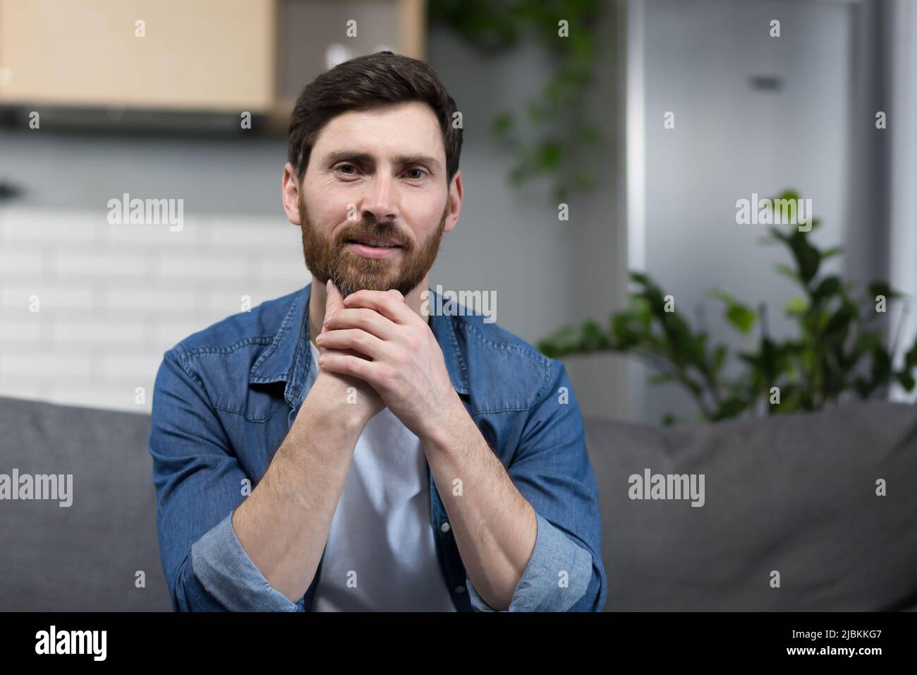 Porträt eines Mannes mit Bart zu Hause, der auf der Couch sitzt und die Kamera anschaut Stockfoto