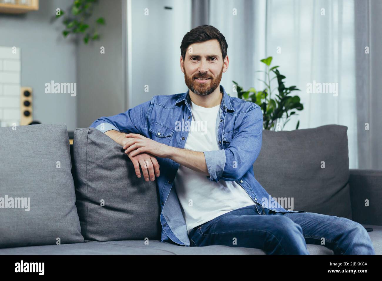 Porträt eines Mannes mit Bart zu Hause, der auf der Couch sitzt und die Kamera anschaut Stockfoto