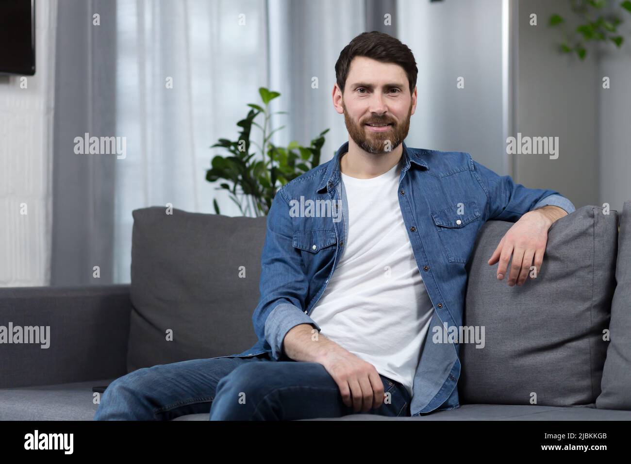 Der Mann zu Hause sitzt auf der Couch, ruht sich aus und schaut weg, lächelt verträumt nachdenklich Stockfoto
