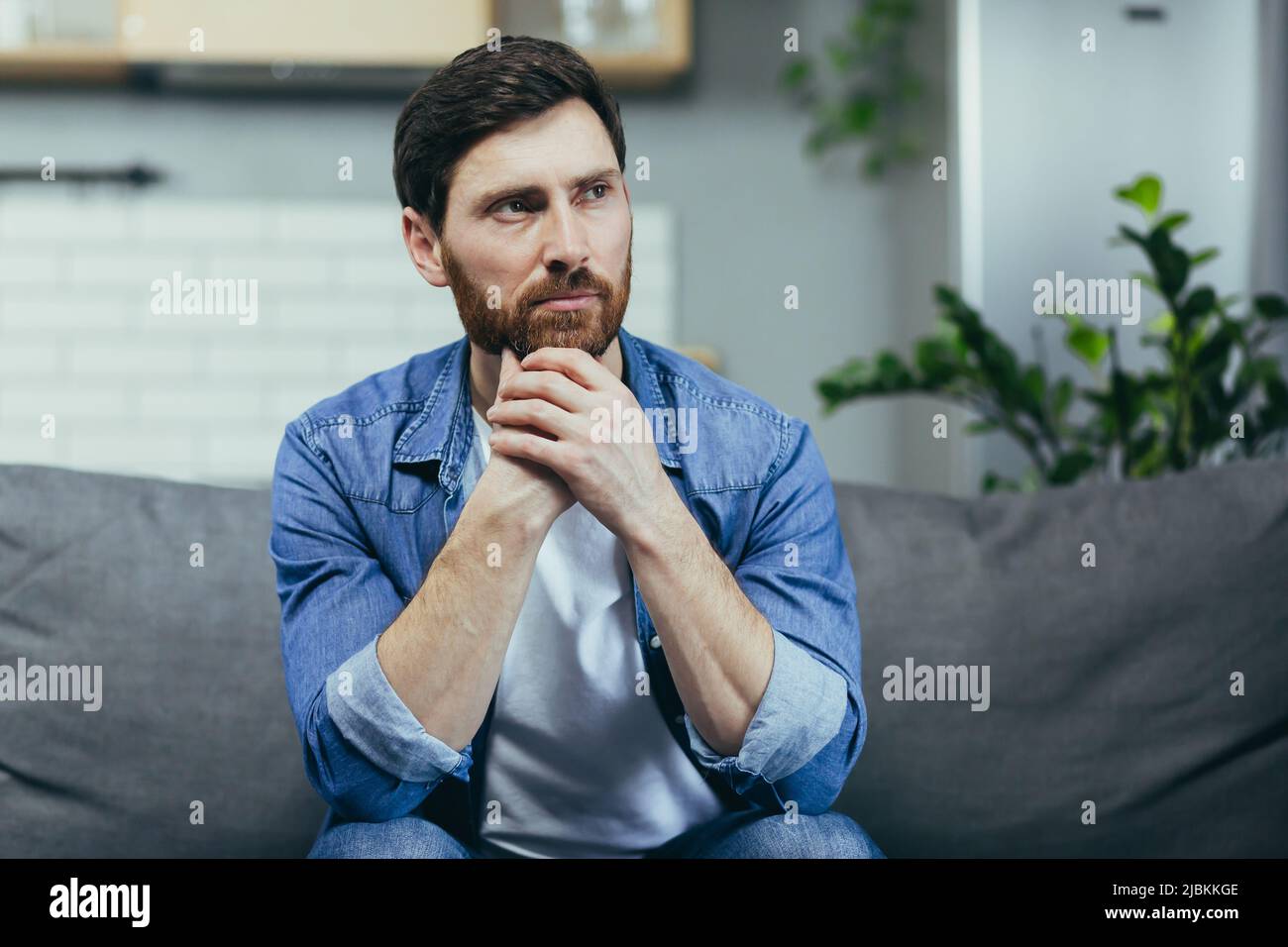 Der Mann zu Hause sitzt auf der Couch, ruht sich aus und schaut weg, lächelt verträumt nachdenklich Stockfoto