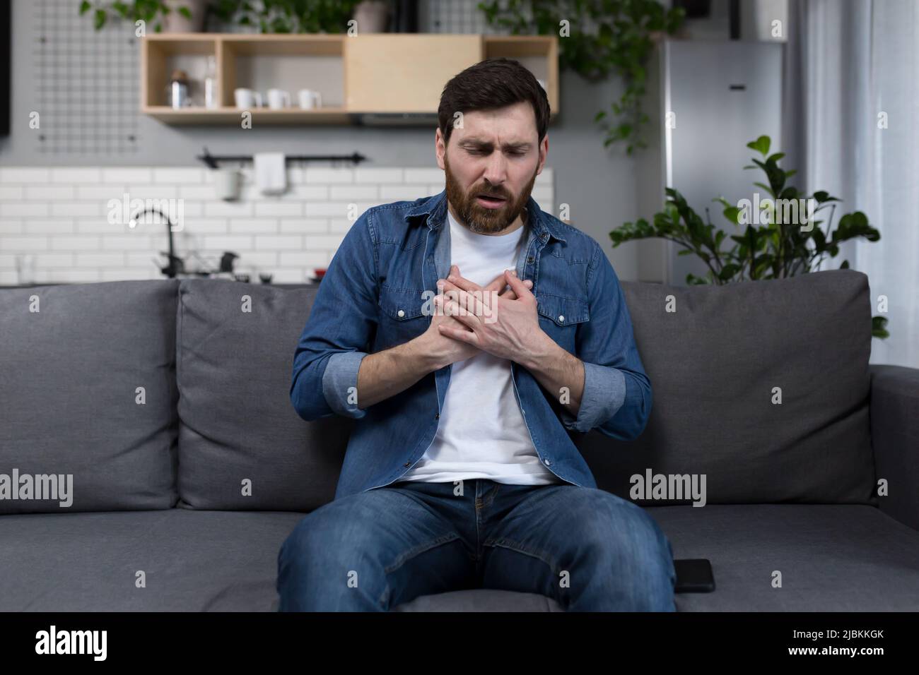Kranker Mann zu Hause, plötzlicher Herzschmerz, sitzt auf der Couch und hält die Hände auf seiner Brust Stockfoto
