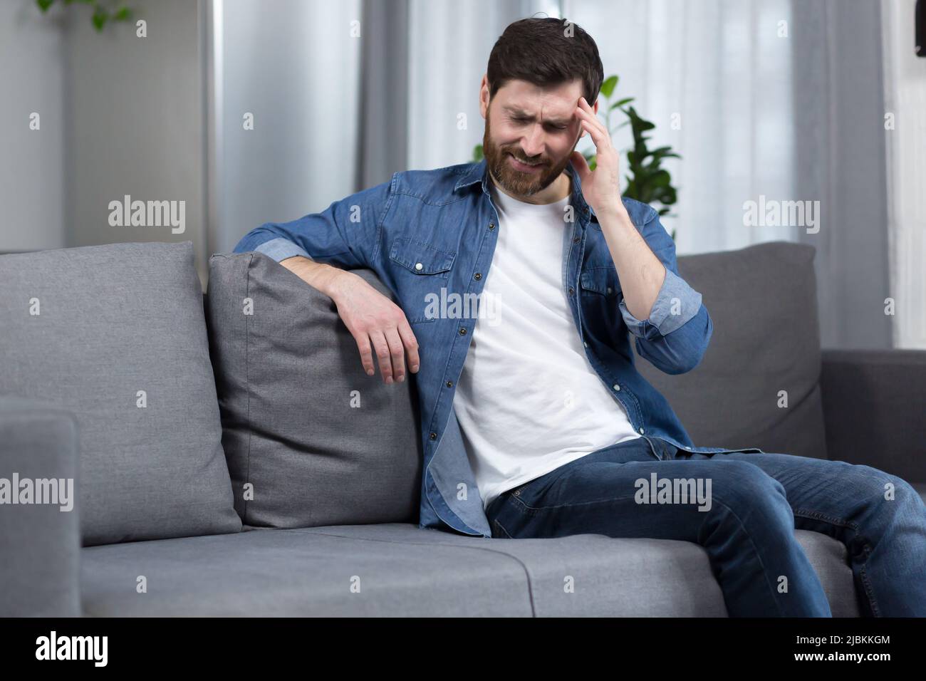 Der Mann ist zu Hause, der Patient sitzt auf der Couch, hat starke Kopfschmerzen Stockfoto