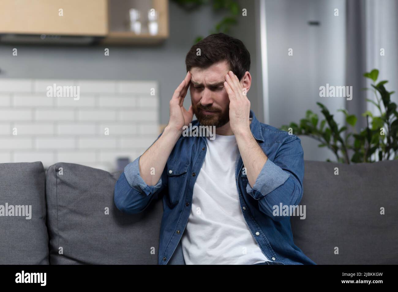 Der Mann ist zu Hause krank, hat starke Kopfschmerzen und sitzt auf der Couch in der Küche Stockfoto