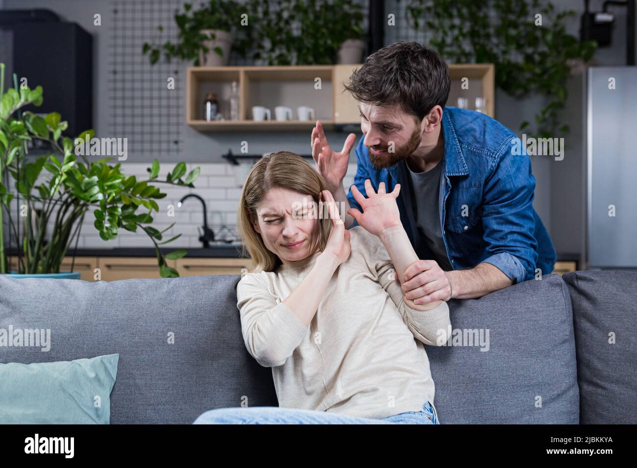 Häusliche Gewalt. Familienkonflikt. Ein Mann schreit und schlägt eine Frau zu Hause. Verängstigte Frau schließt ihre Arme und sitzt auf der Couch. Sie hat Angst Stockfoto