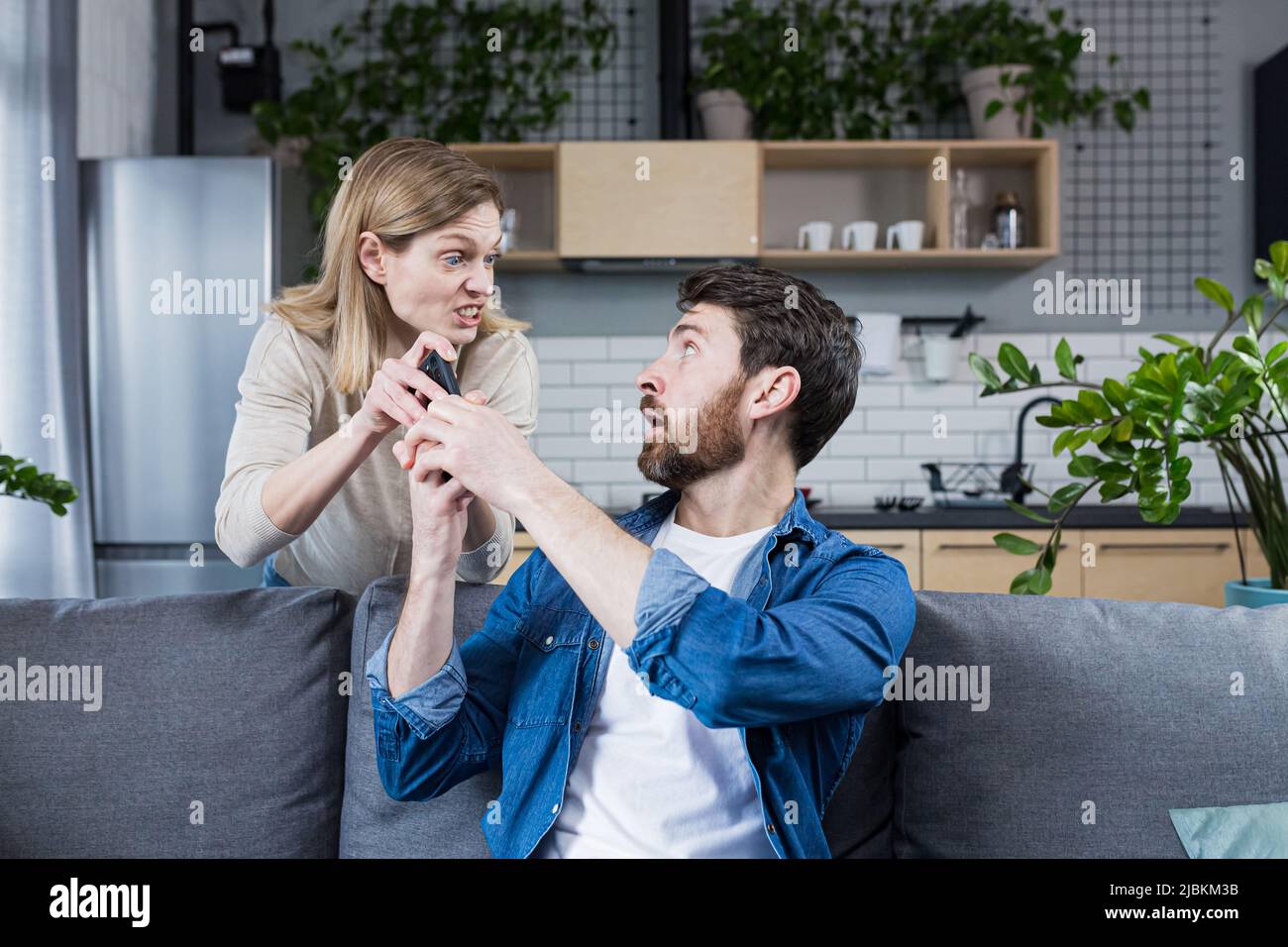 Konflikt in der Familie. Die Frau einer wütenden, verärgerten Frau nimmt das Telefon des Mannes, der zu Hause auf der Couch sitzt, und nutzt das Telefon ständig. Stockfoto