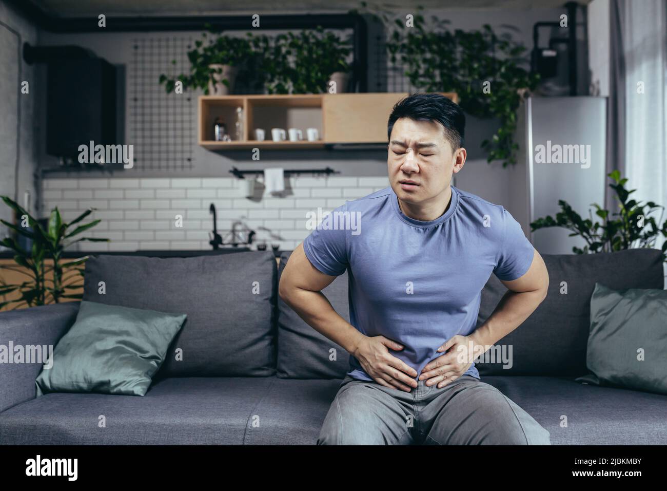 Asiatischer Mann selbst zu Hause krank auf der Couch sitzen, hat starke Bauchschmerzen, Mann im Wohnzimmer Stockfoto