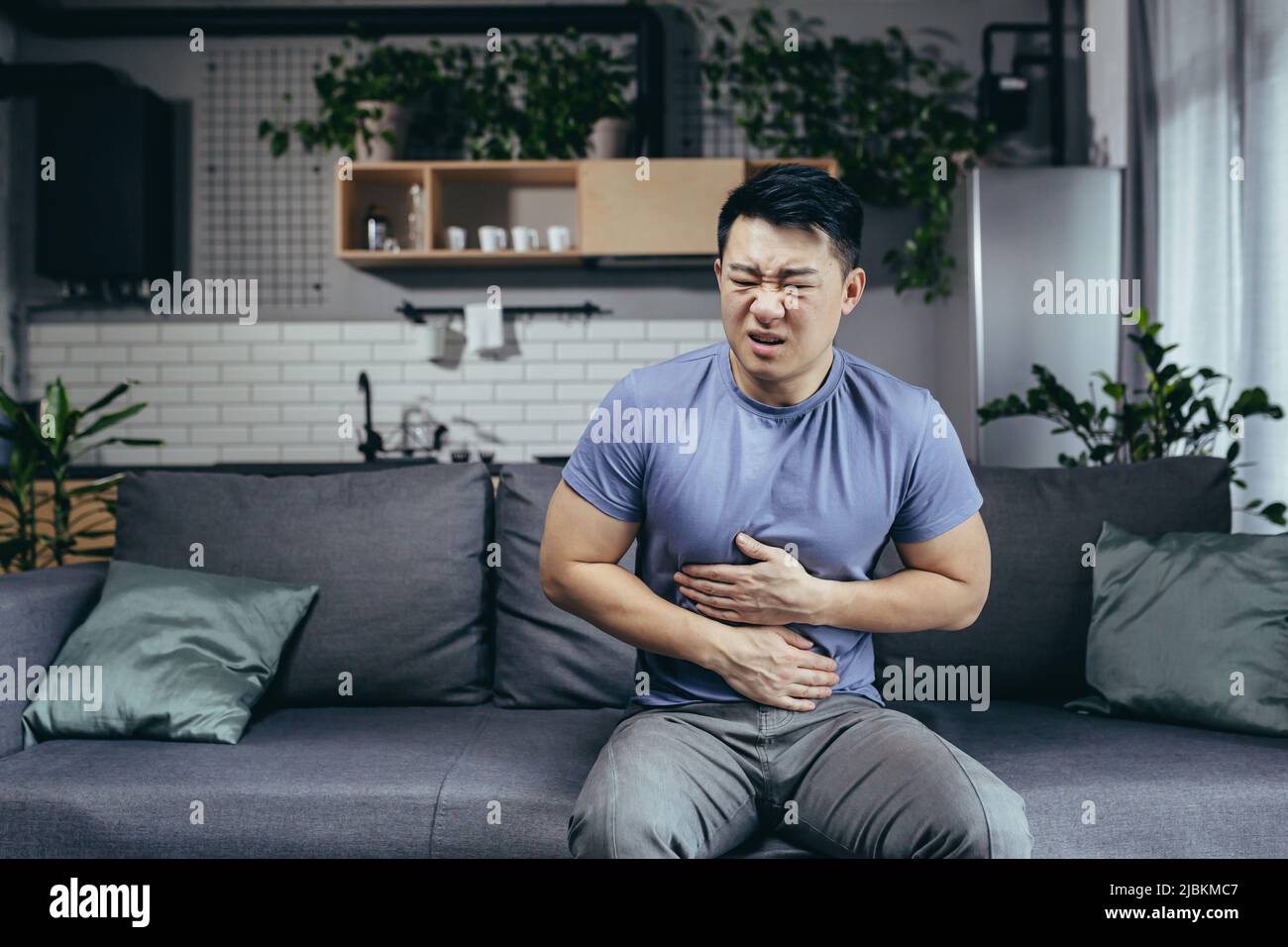 Asiatischer Mann selbst zu Hause krank auf der Couch sitzen, hat starke Bauchschmerzen, Mann im Wohnzimmer Stockfoto