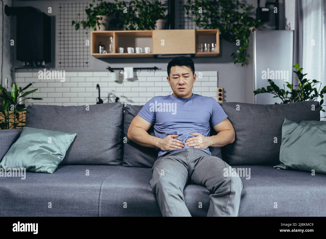Asiatischer Mann selbst zu Hause krank auf der Couch sitzen, hat starke Bauchschmerzen, Mann im Wohnzimmer Stockfoto