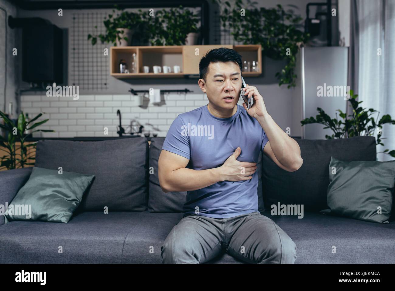 Der asiatische Mann allein zu Hause, hat starke Brustschmerzen, ruft einen Arzt, meldet seine Krankheit, sitzt zu Hause auf der Couch Stockfoto