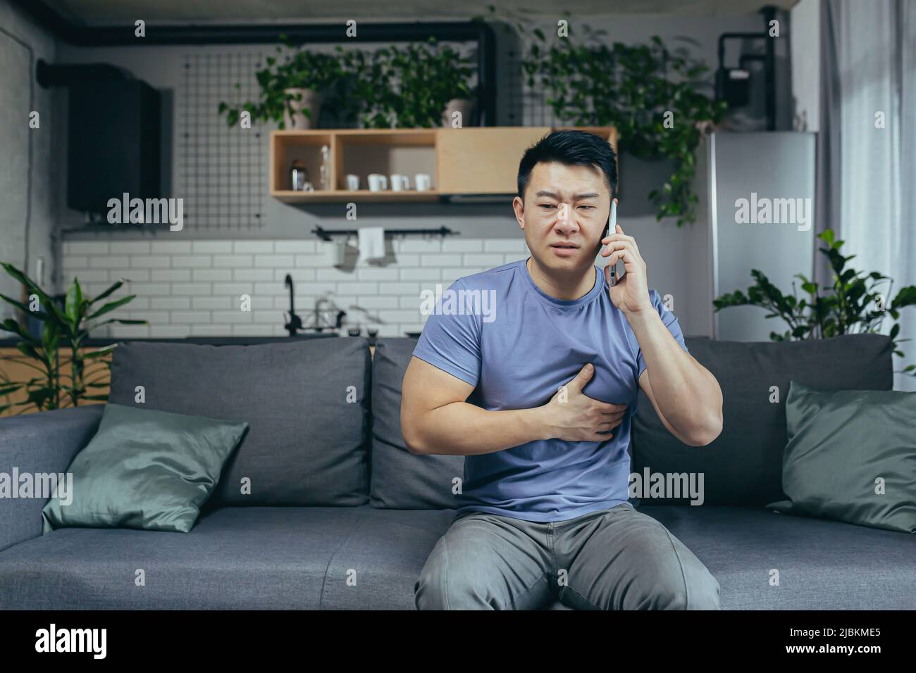 Der asiatische Mann allein zu Hause, hat starke Brustschmerzen, ruft einen Arzt, meldet seine Krankheit, sitzt zu Hause auf der Couch Stockfoto