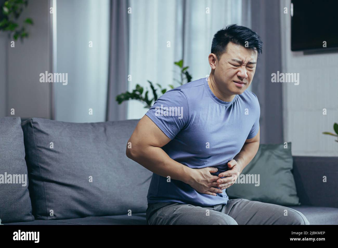 Asiatischer Mann selbst zu Hause krank auf der Couch sitzen, hat starke Bauchschmerzen, Mann im Wohnzimmer Stockfoto