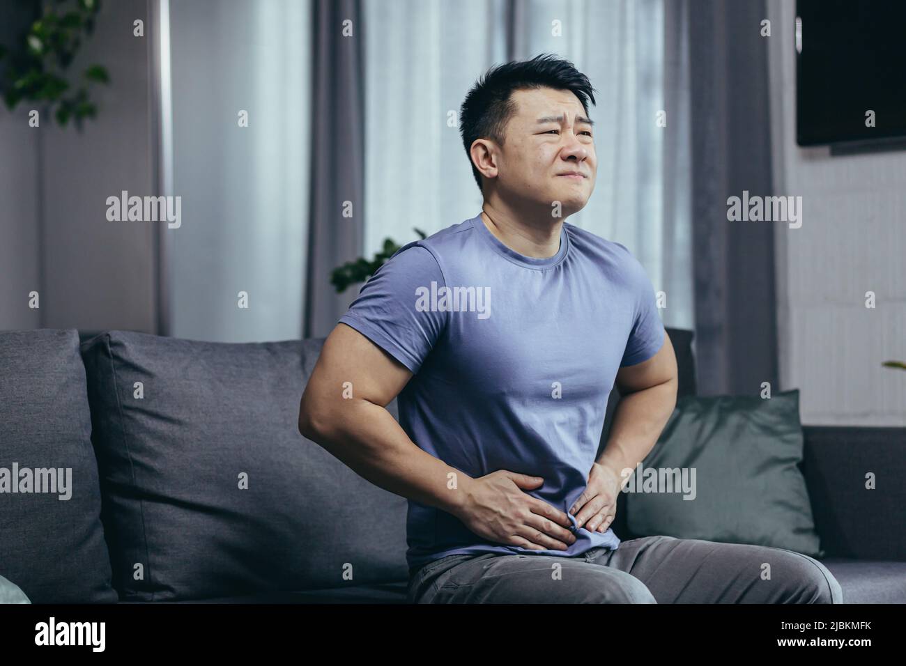 Asiatischer Mann selbst zu Hause krank auf der Couch sitzen, hat starke Bauchschmerzen, Mann im Wohnzimmer Stockfoto