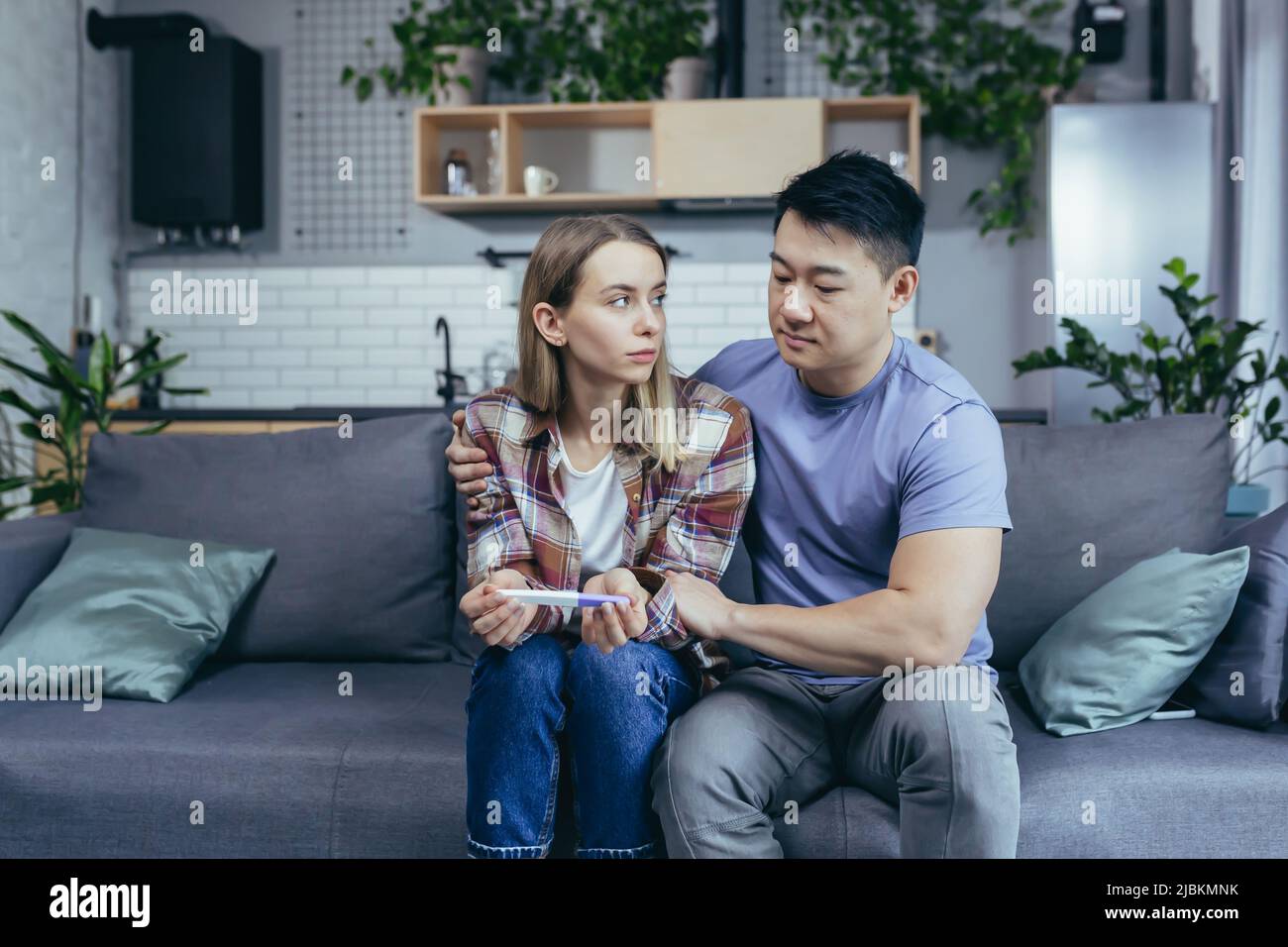 Mann Asian unterstützt Frau, enttäuschende Schwangerschaftstest Ergebnis, junge multirassische Paar sitzt auf dem Sofa, frustriert Stockfoto