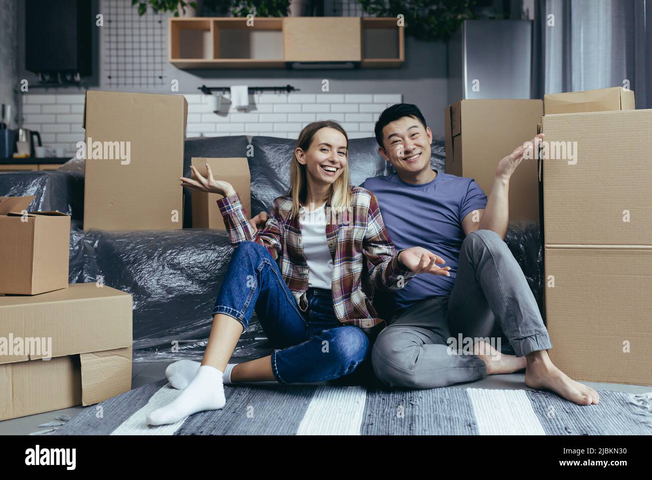 Junge, multirassische Familie, Mann und Frau, die zwischen Kartons auf dem Boden sitzen, auf die Kamera schauen und lächelnde, winkende Hände zum Gruß bewegen Stockfoto
