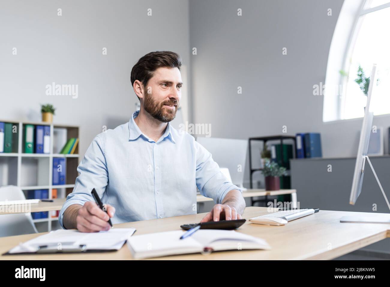 Porträt eines jungen Mannes. Buchhalter, Ökonom, Finanzierer. Setzt auf einen Rechner, sitzt am Schreibtisch am Computer, arbeitet mit Dokumenten Stockfoto