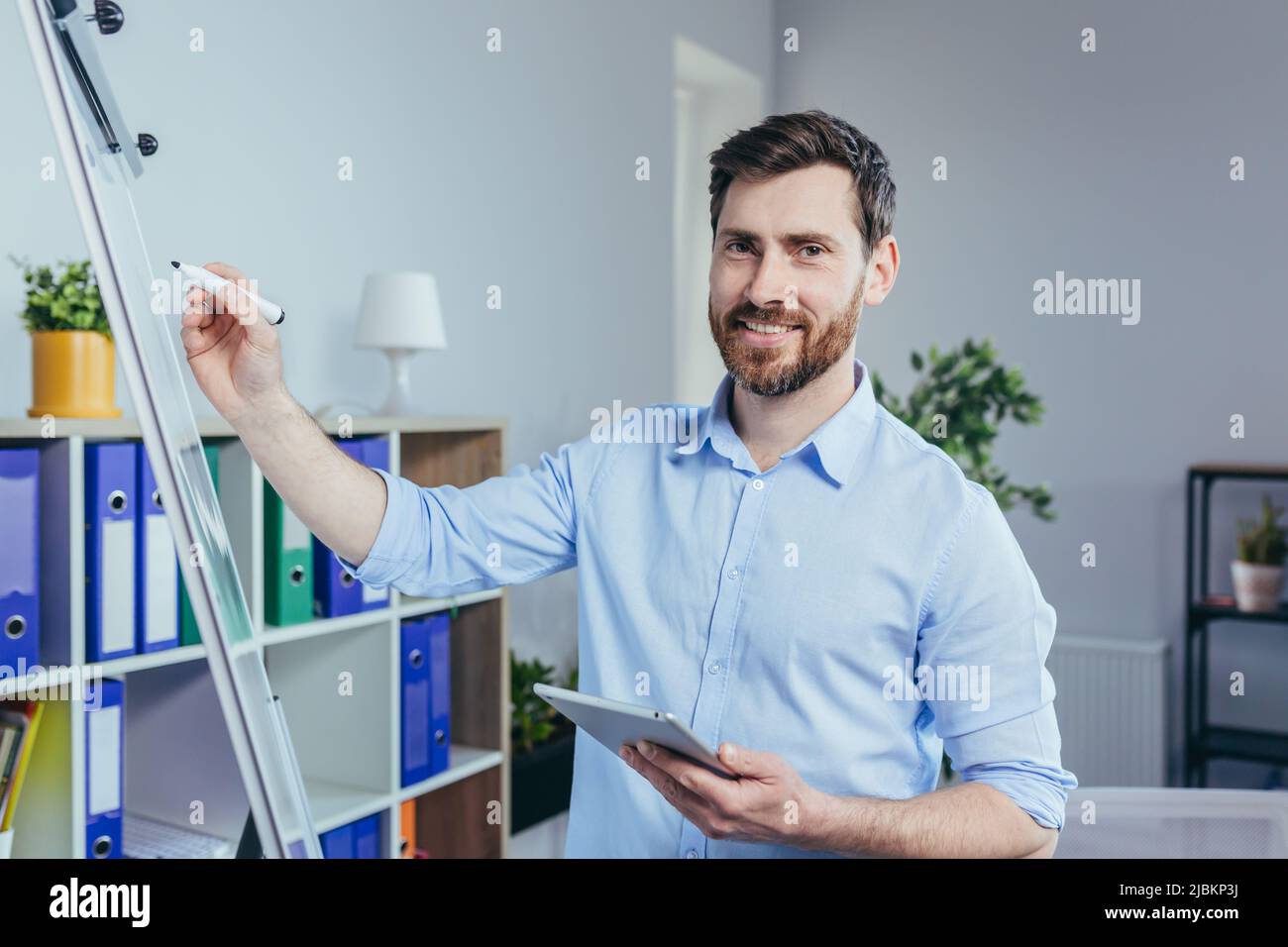 Porträt eines erfolgreichen Unternehmensgründers, ein Mann schaut auf die Kamera und lächelt auf ein Whiteboard für Notizen, hält ein Tablet, beschreibt eine Business str Stockfoto