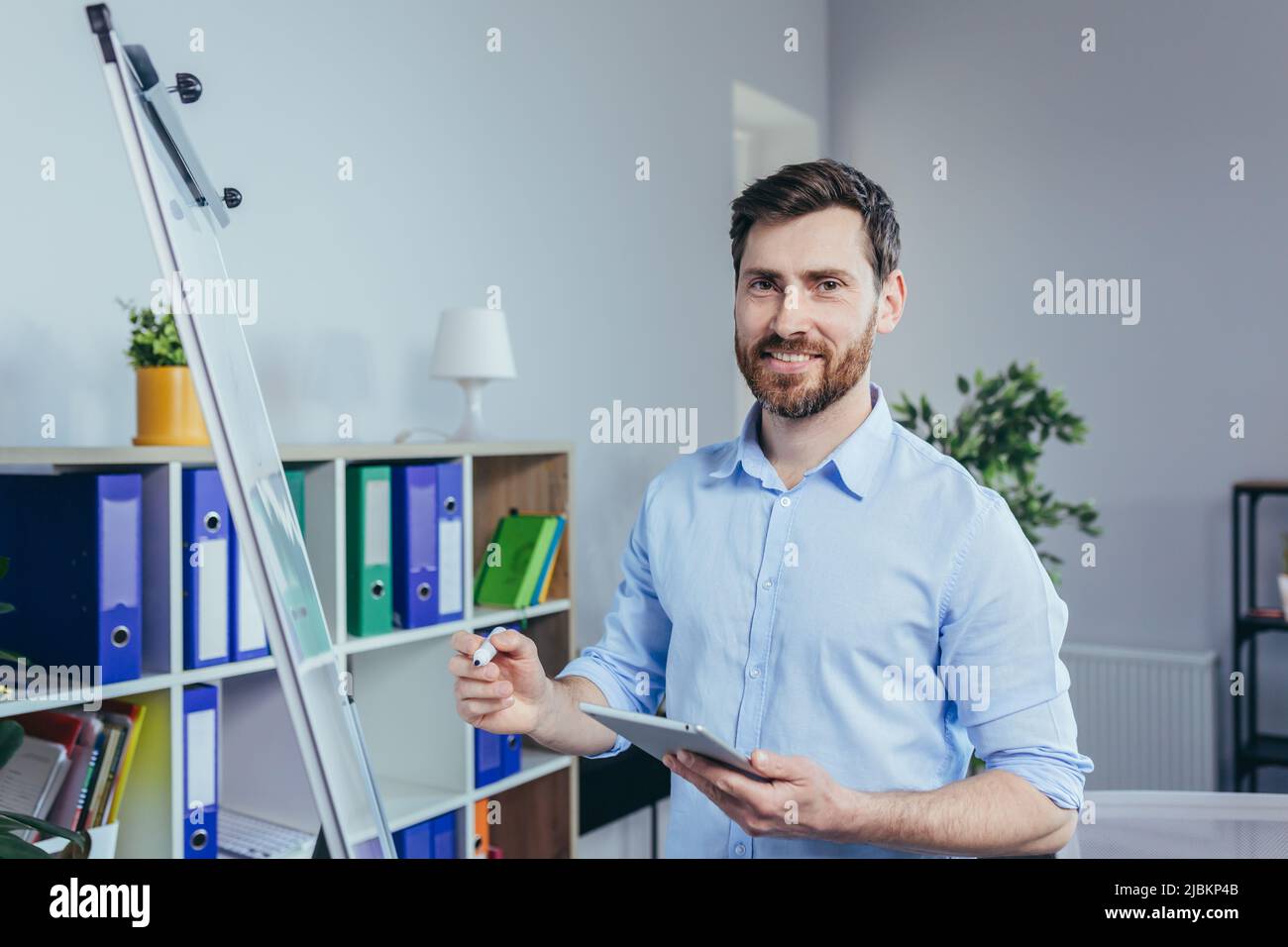 Porträt eines erfolgreichen Unternehmensgründers, ein Mann schaut auf die Kamera und lächelt auf ein Whiteboard für Notizen, hält ein Tablet, beschreibt eine Business str Stockfoto