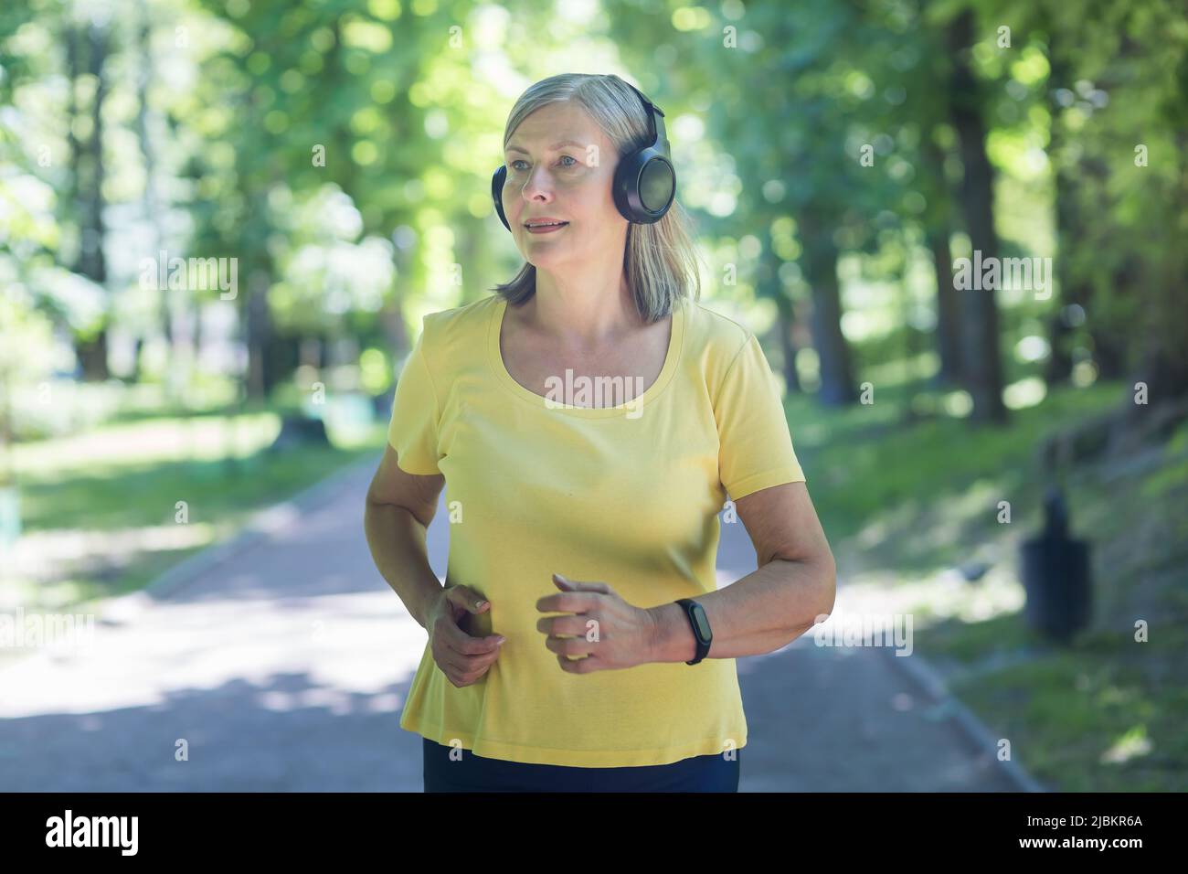 Ältere schöne Frau mit Kopfhörern, die im Park läuft, morgens joggt, aktiv ist, Musik hört. Fühlt sich gut an, lächelt Stockfoto