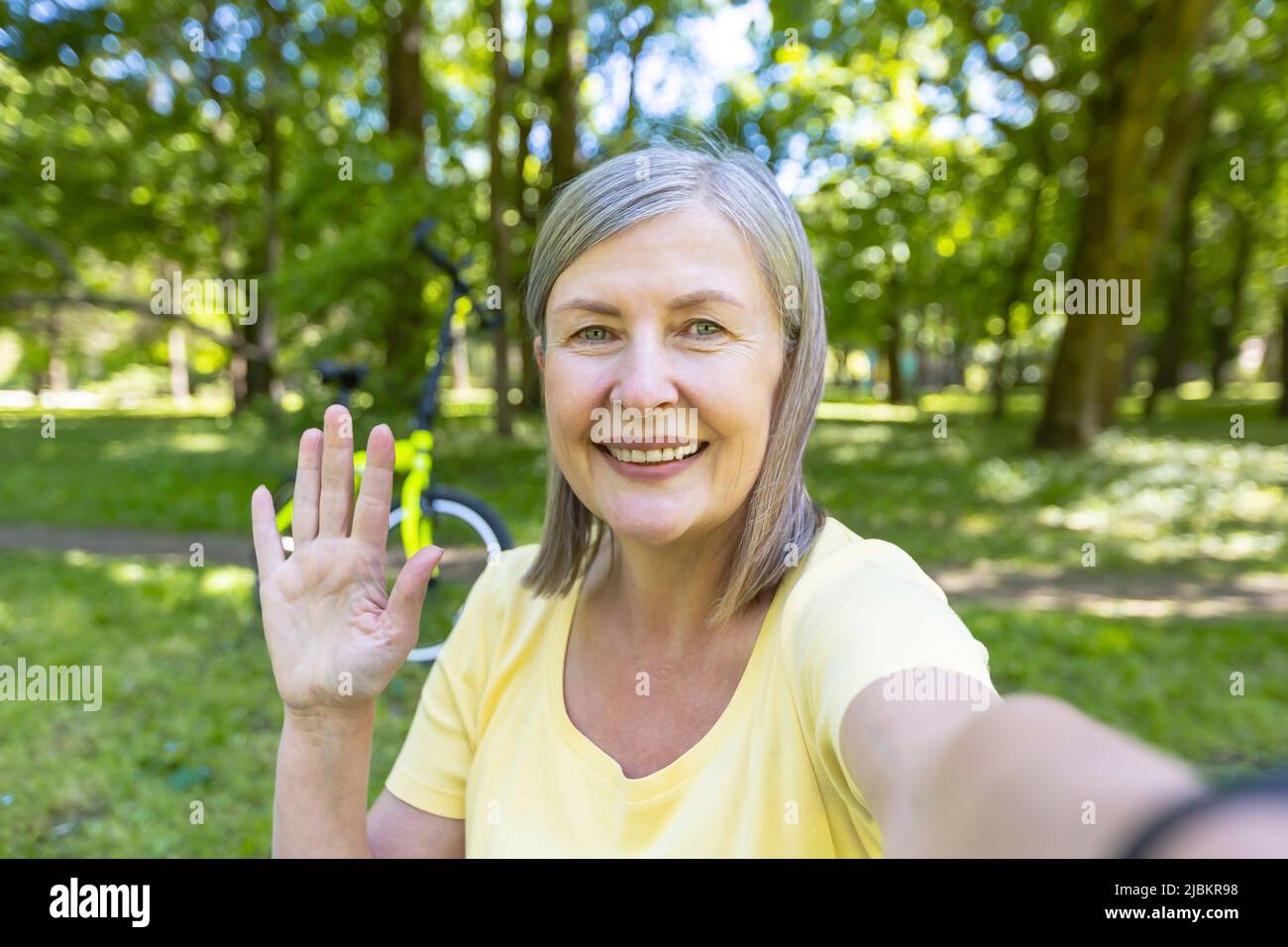 Ältere grauhaarige Frau, aktive Sportlerin im Park im Sommer blickt auf die Telefonkamera und lächelt mit der Hand auf einen Videoanruf Stockfoto