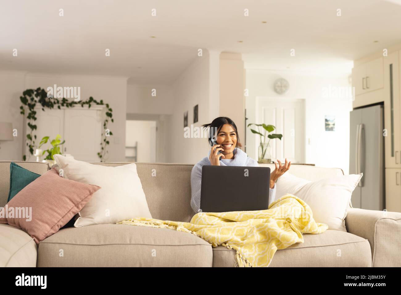 Lächelnde junge Birazialin, die auf einem Smartphone spricht, während sie zu Hause mit einem Laptop sitzt Stockfoto