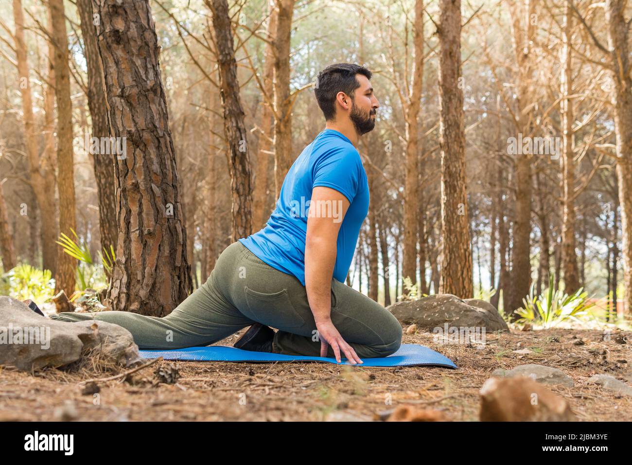 Hübscher junger Mann, der bei Sonnenuntergang im Wald, umgeben von Bäumen auf seiner blauen Matte, Yoga-Dehnung praktiziert. Stockfoto