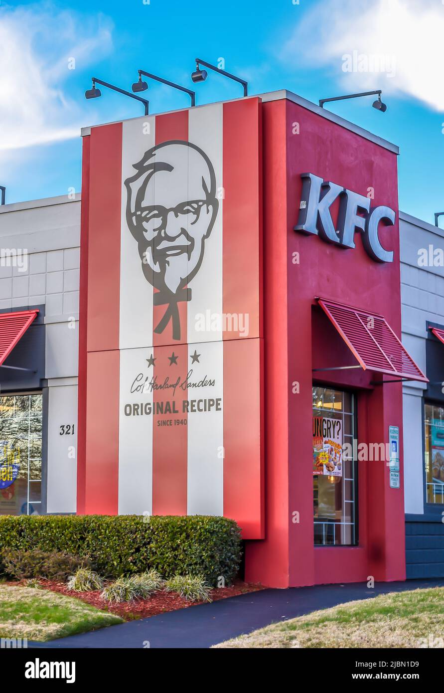 Die Außenfassade des KFC Fast Food-Restaurants mit frittierten Hühnchen und das Logo an einem hellen, sonnigen Tag mit Sträuchern, blauem Himmel und Außenbeleuchtung. Stockfoto