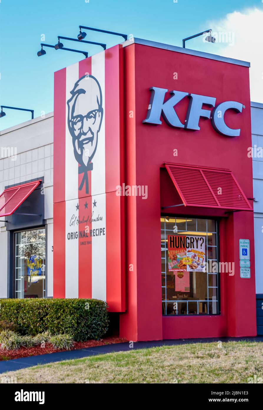 Die Außenfassade des KFC Fast Food-Restaurants mit frittierten Hühnchen und das Logo an einem hellen, sonnigen Tag mit Sträuchern, blauem Himmel und Außenbeleuchtung. Stockfoto