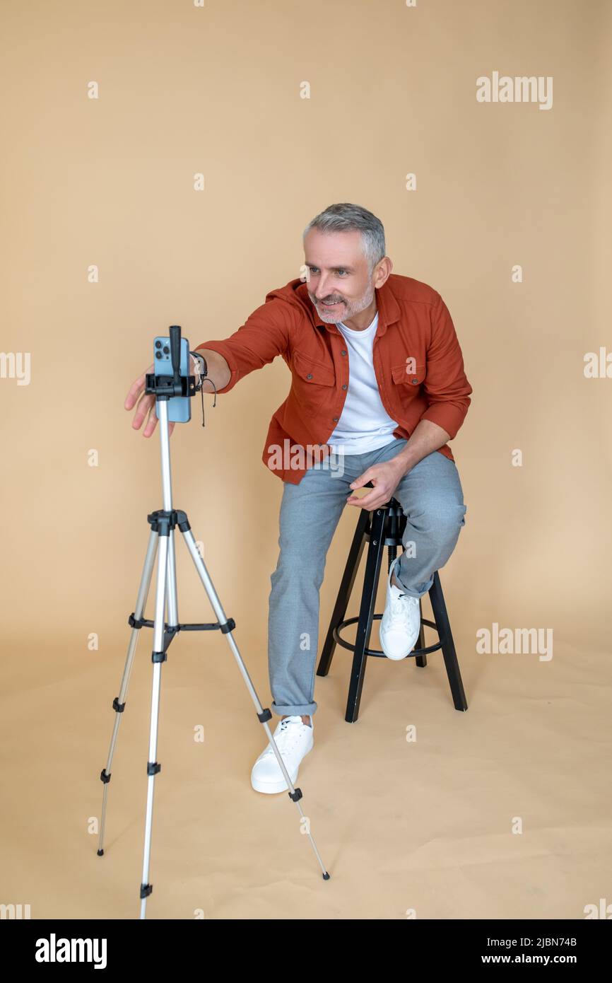 Ein Mann in einer roten Jacke, der in einem Studio auf einem Hocker sitzt und Selfie macht Stockfoto