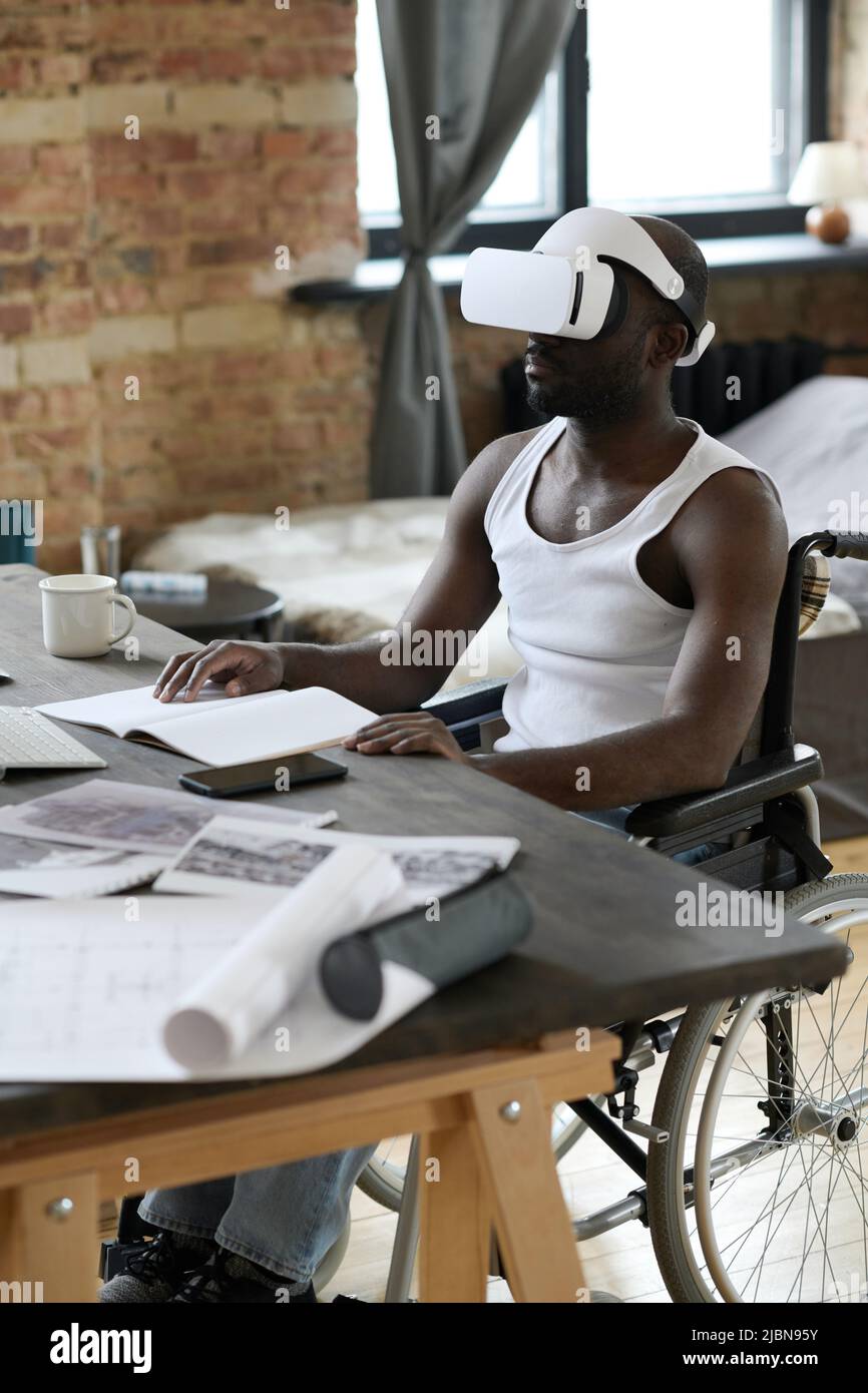 Afroamerikanischer Mann, der im Rollstuhl am Schreibtisch sitzt und dabei eine Virtual-Reality-Brille verwendet, um Architekturprojekte zu lernen Stockfoto