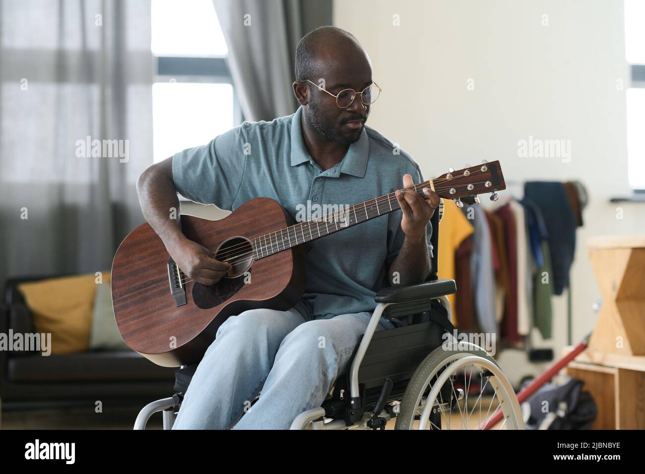 Afroamerikanischer Mann in Brillen, der im Rollstuhl sitzt und in seiner Freizeit zu Hause Gitarre spielen lernt Stockfoto