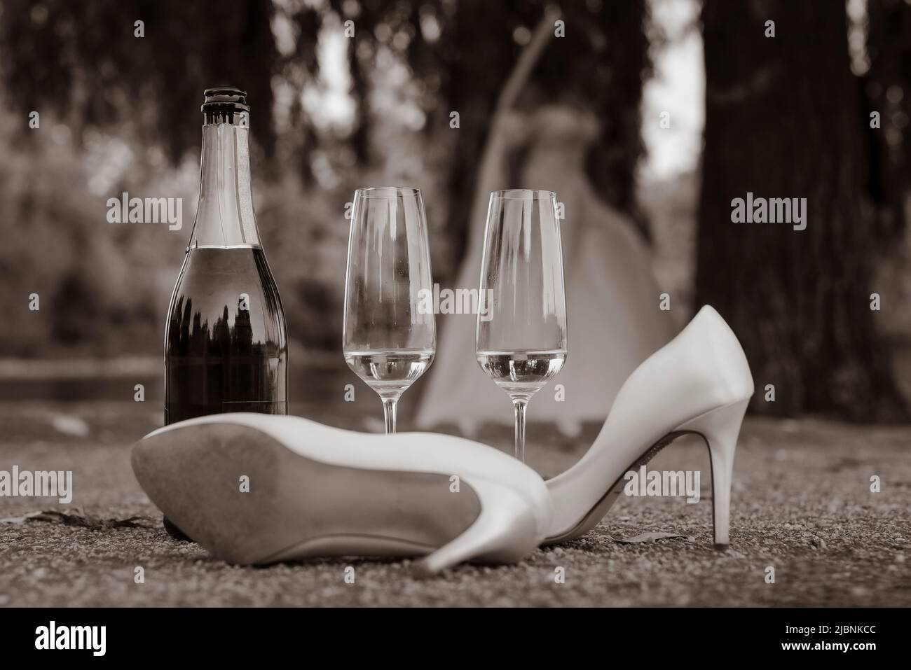 Eine Champagnerflasche, ein Paar High Heels und zwei Sektgläser befinden sich auf dem Boden im Freien, Ende einer Hochzeit, Schwarz-Weiß-Foto Stockfoto