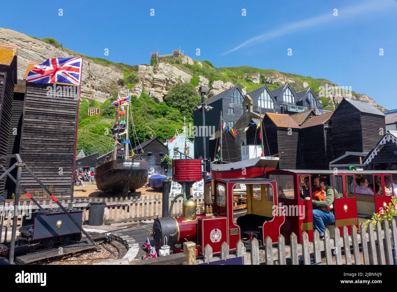 Menschen an Bord eines Miniatureisenbahnzuges an der Küste von Hastings, mit einem East Hill Cliff Lift im Hintergrund, East Sussex, Großbritannien Stockfoto