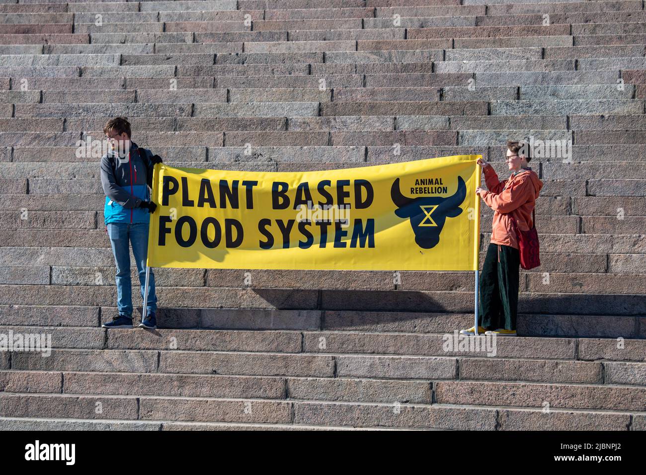 Zwei Personen mit einem gelben Banner, das ein pflanzenbasiertes Ernährungssystem auf der Helsinki Cathdedral Steps in Helsinki, Finnland, fordert Stockfoto