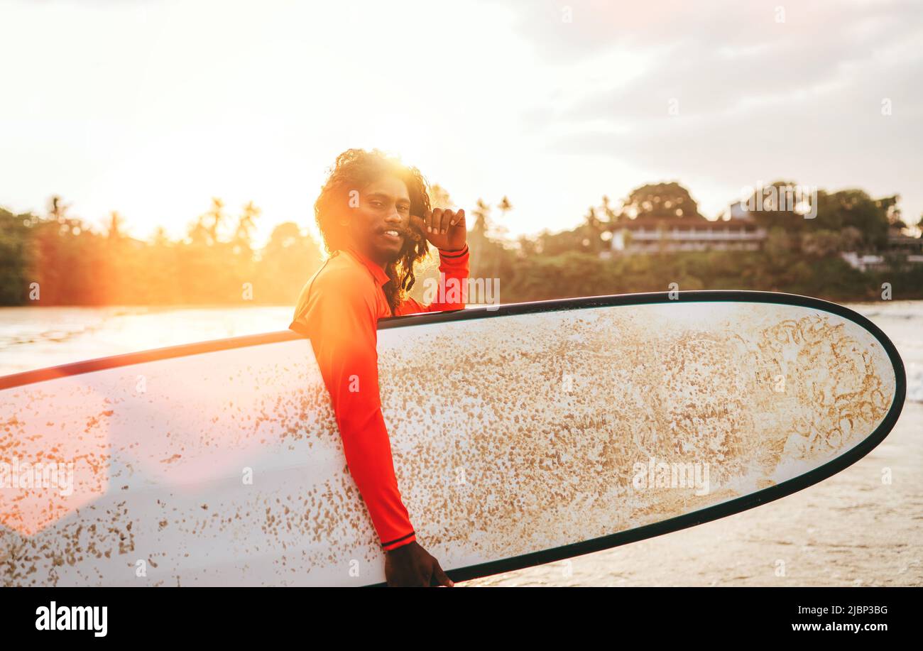 Porträt eines schwarzen langhaarigen Teenagers mit einem Surfbrett, das mit Sonnenuntergang-Hintergrundbeleuchtung zum Surfen bereit ist. Er läuft in die Wellen des indischen Ozeans. spo bei extremem Wasser Stockfoto