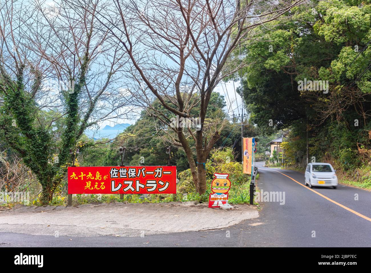 kyushu, japan - 09 2021. dezember: Tafel und Maskottchen neben der Straße, die zu einem berühmten Sasebo Burger Restaurant im Saikai Nationalpark führt, mit Blick auf die Stockfoto