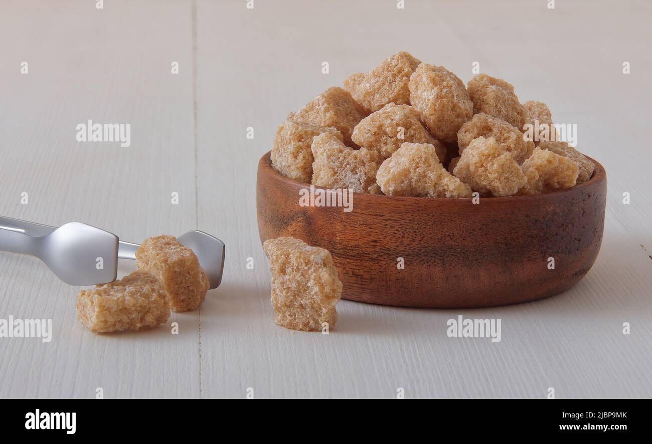 Würfelförmiger Rohrzucker in Holzschüssel auf einem weißen Holztisch mit silberner Zuckerzange. Low-Angle-Ansicht, keine Menschen. Stockfoto